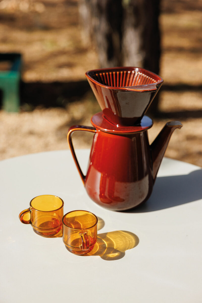 Cafetière en céramique brun roux et deux tasses en verre ambré posées sur une table blanche, en plein soleil
