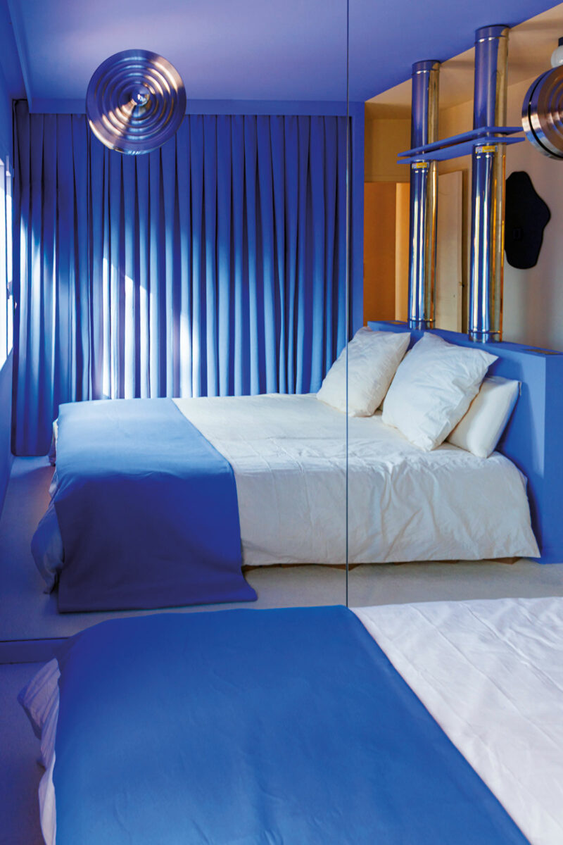 Chambre bleu Ultra-Marin avec rideaux plissés, suspension argentée et conduits métalliques en tête de lit