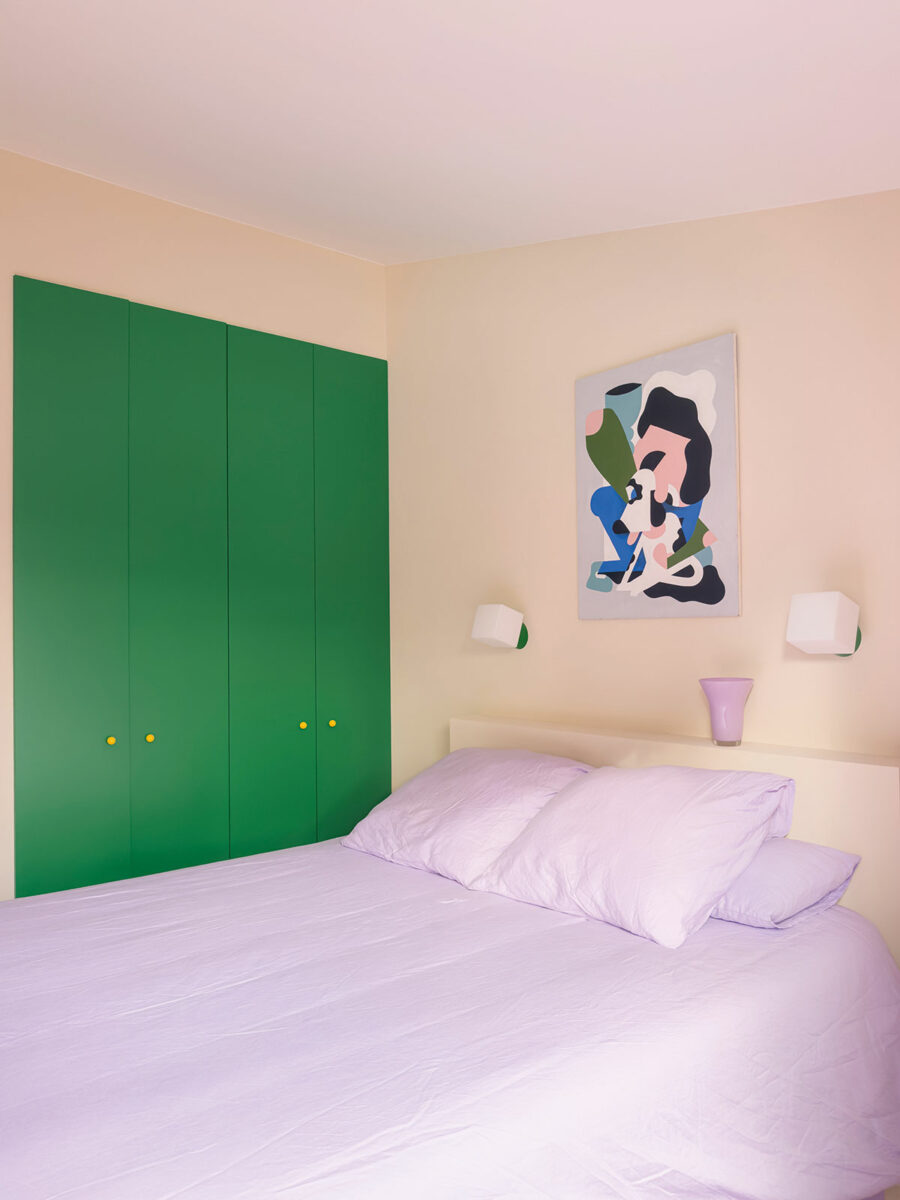 Chambre aux murs rose pâle, linge de lit parme et grand placard vert aux boutons jaunes, sous une toile abstraite