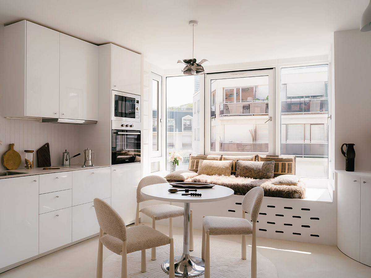 Cuisine blanche ouverte sur le séjour, avec table ronde, chaises en bouclé et banquette sous la fenêtre
