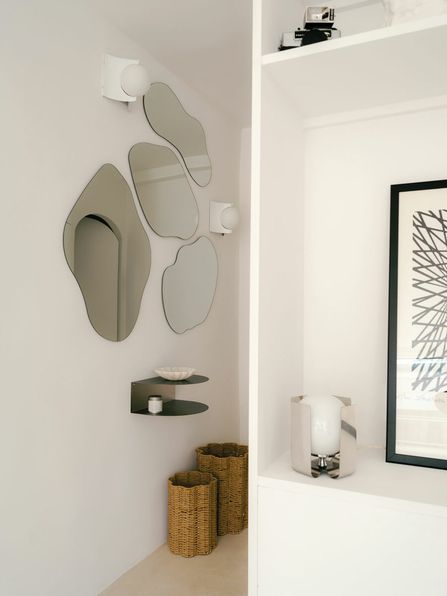 Mur de miroirs aux formes organiques, appliques à globe blanc, étagère en métal et luminaire en inox poli près d’une estampe encadrée