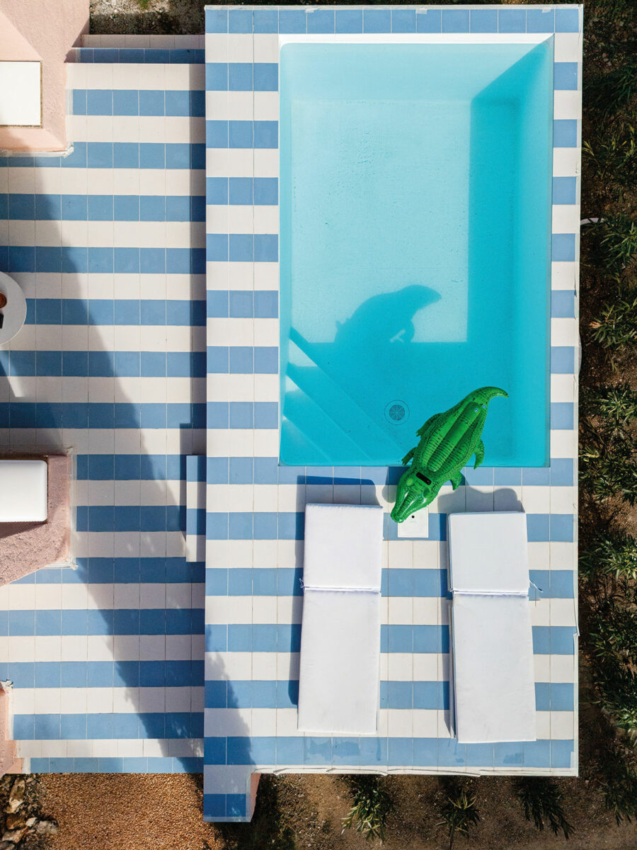 Vue du ciel de la piscine hors sol et de sa terrasse aux larges rayures bleu ciel et blanches, avec une bouée crocodile verte