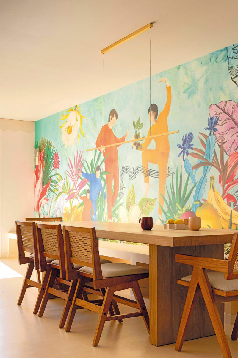 Grande table en bois et chaises cannées devant la fresque murale aux personnages et feuillages colorés