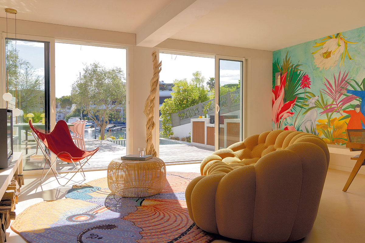 Salon ouvert sur la terrasse et le canal, avec fresque murale, tapis coloré et canapé aux formes rondes