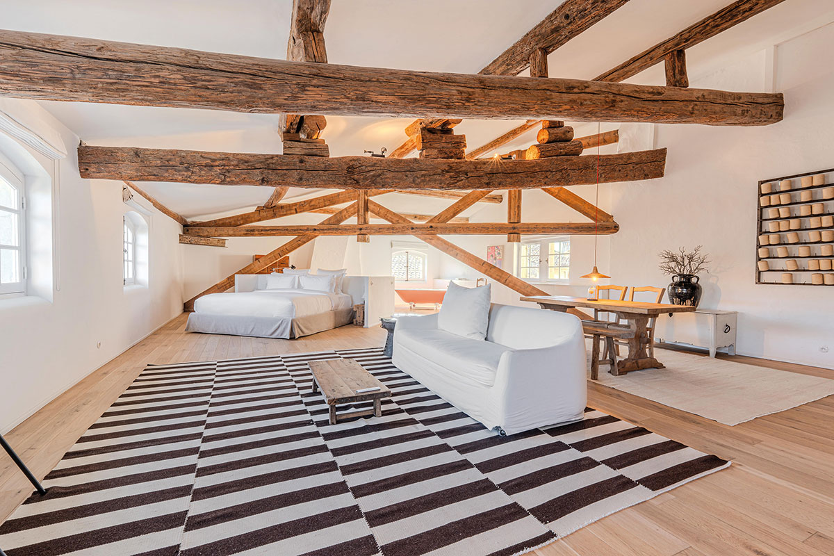 Suite sous charpente aux grandes poutres anciennes, lit, canapé blanc, tapis rayé noir et blanc et baignoire orangée