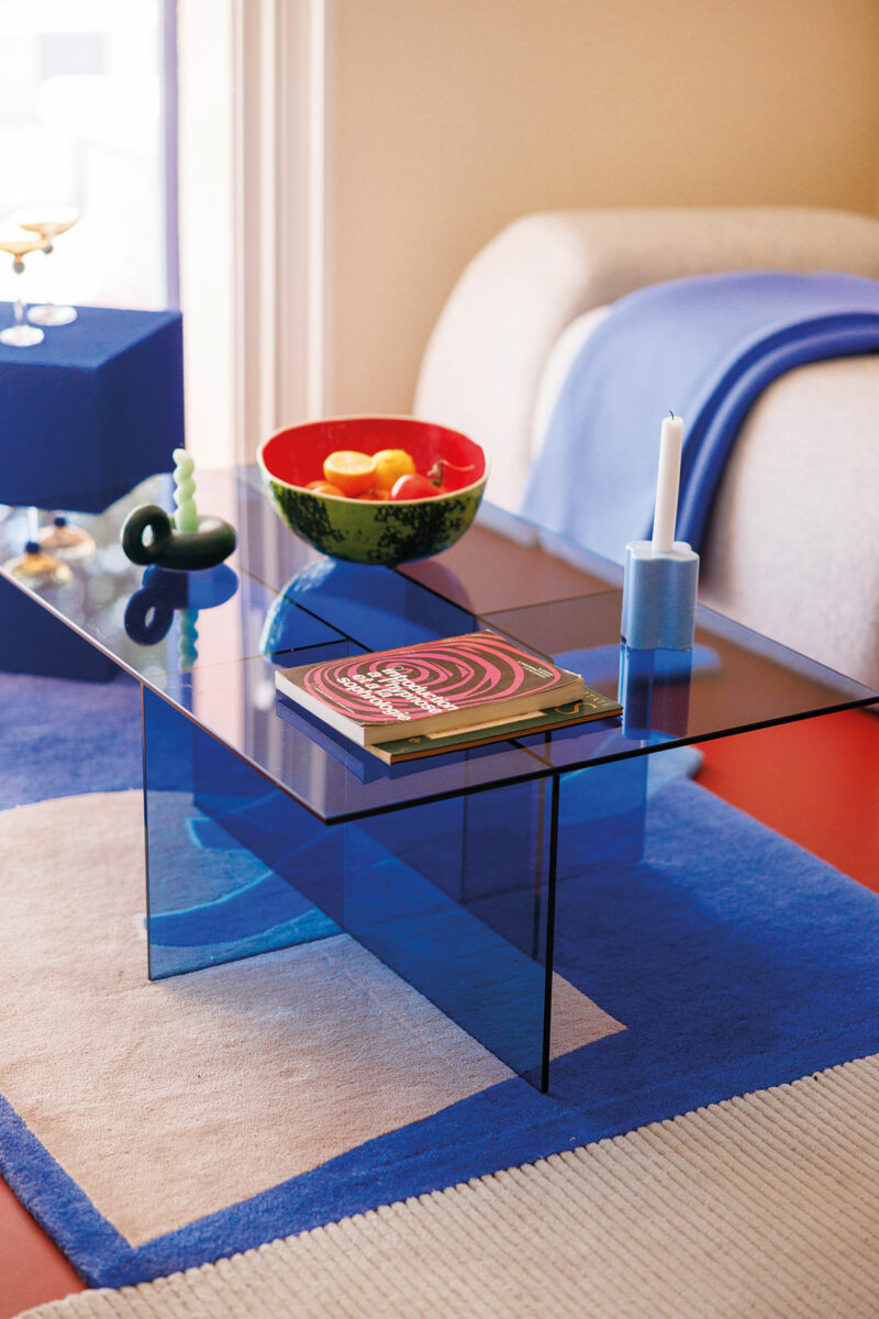 Table basse en verre bleu posée sur un tapis graphique bleu et beige, bol en forme de pastèque et bougeoirs colorés