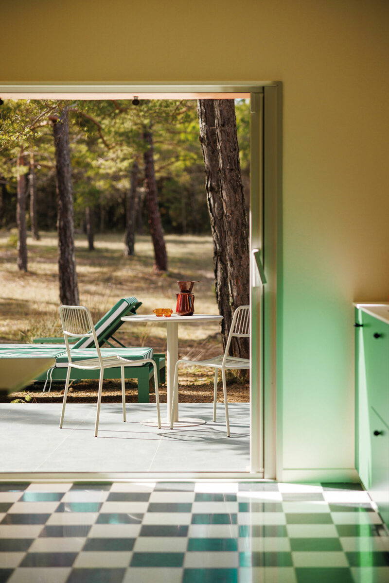 Terrasse ombragée par les pins, vue depuis la cuisine à travers la baie ouverte, avec table et chaises blanches et transats verts