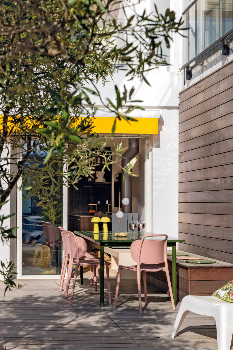Terrasse ombragée par l’olivier, avec table carrelée verte, chaises roses et lampes jaunes devant la cuisine